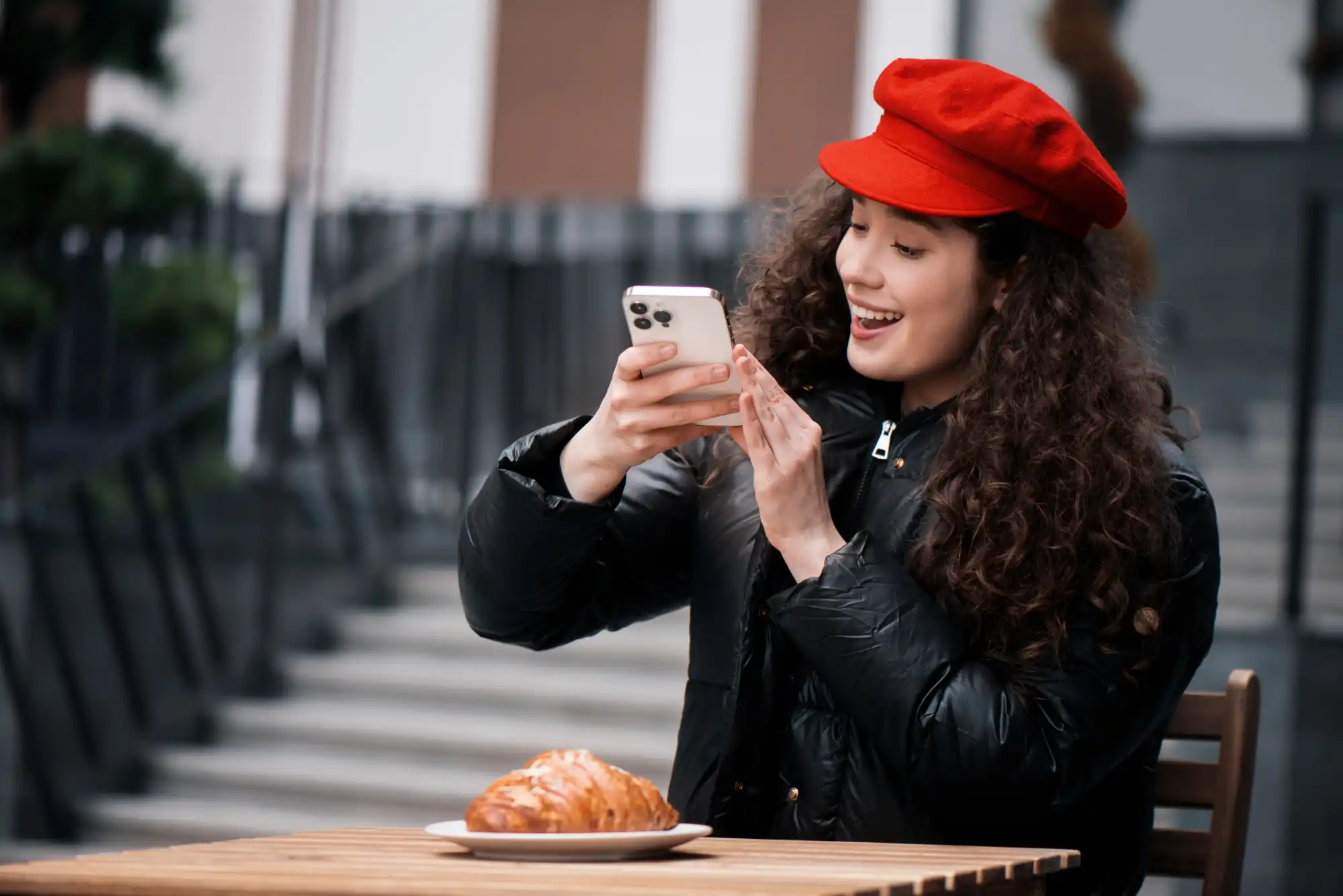 Young French woman in red beret using smartphone for TikTok Shop while sitting at cafe with croissant