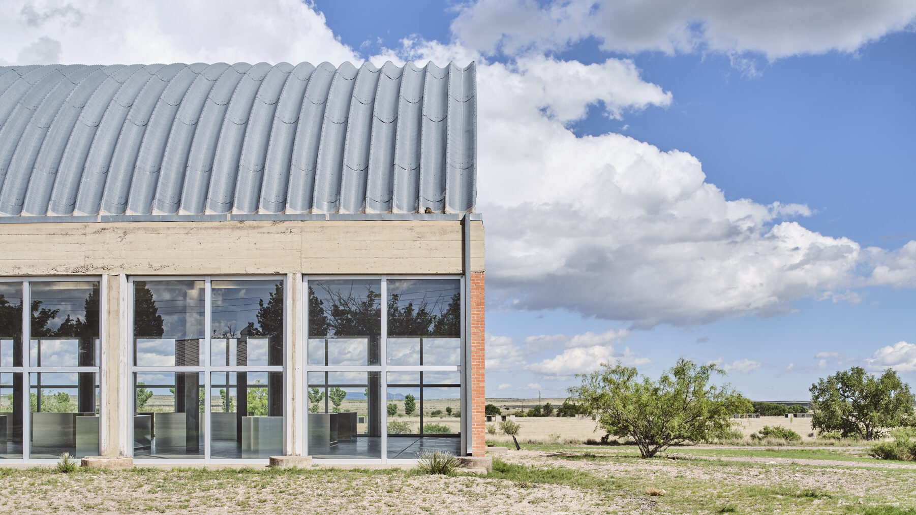Exterior of the Chinati Foundation building in Marfa, Texas, with ribbed metal roof, large glass windows, and open desert landscape under a wide blue sky.