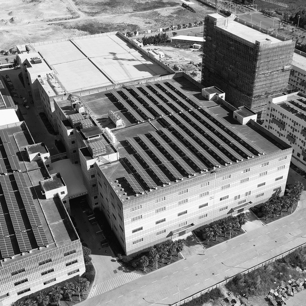 High-angle aerial view of a modern industrial facility and office buildings in a grayscale cityscape. The flat roofs are equipped with extensive solar panel arrays, showcasing sustainable energy infrastructure and industrial architecture.