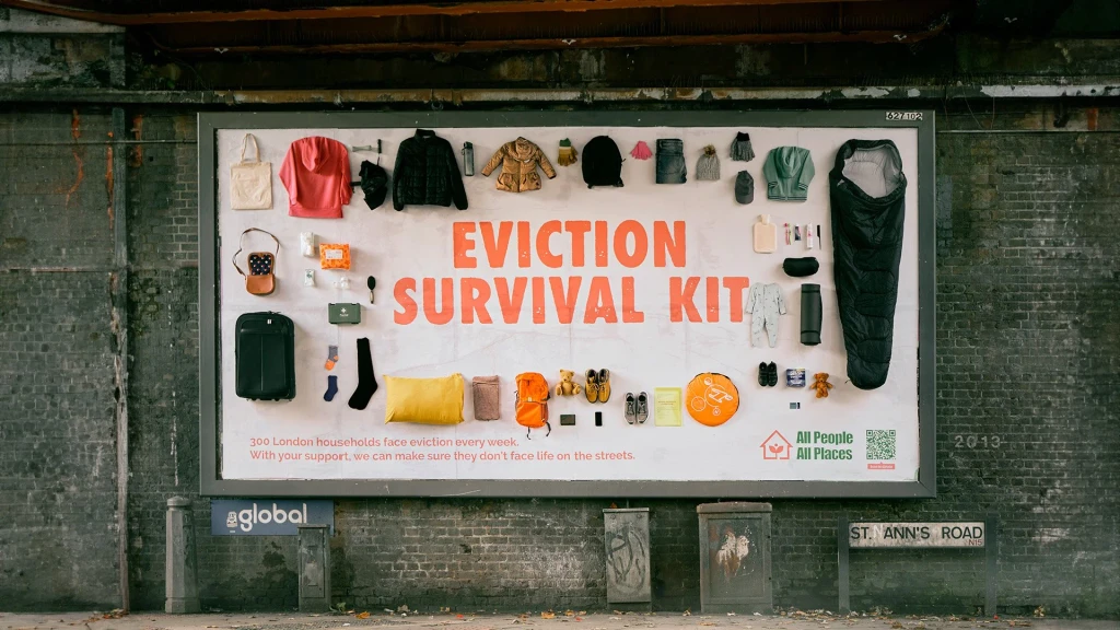 Billboard on a road in London that says Evicition Survival Kit. It has real items like a jumper, sleeping bacg and suitcast attached to it.
