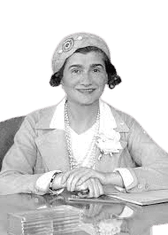 A black-and-white portrait of a smiling woman dressed elegantly in a light-colored suit and a decorative hat. She is seated at a desk with her hands resting calmly in front of her, wearing a pearl necklace and a floral accessory on her outfit. The image captures a poised and confident demeanor, with the soft background drawing focus to her cheerful expression and refined style.