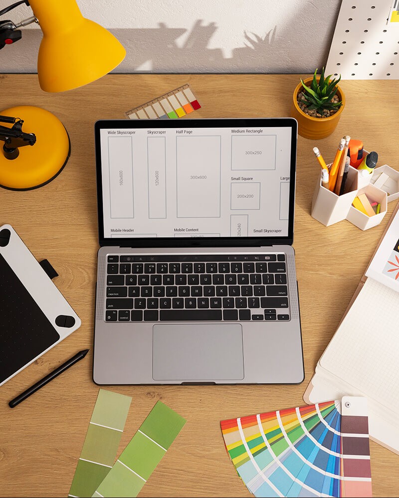 A top-down view of a MacBook with screen showing on a desk, with general graphic designer items scattered around it