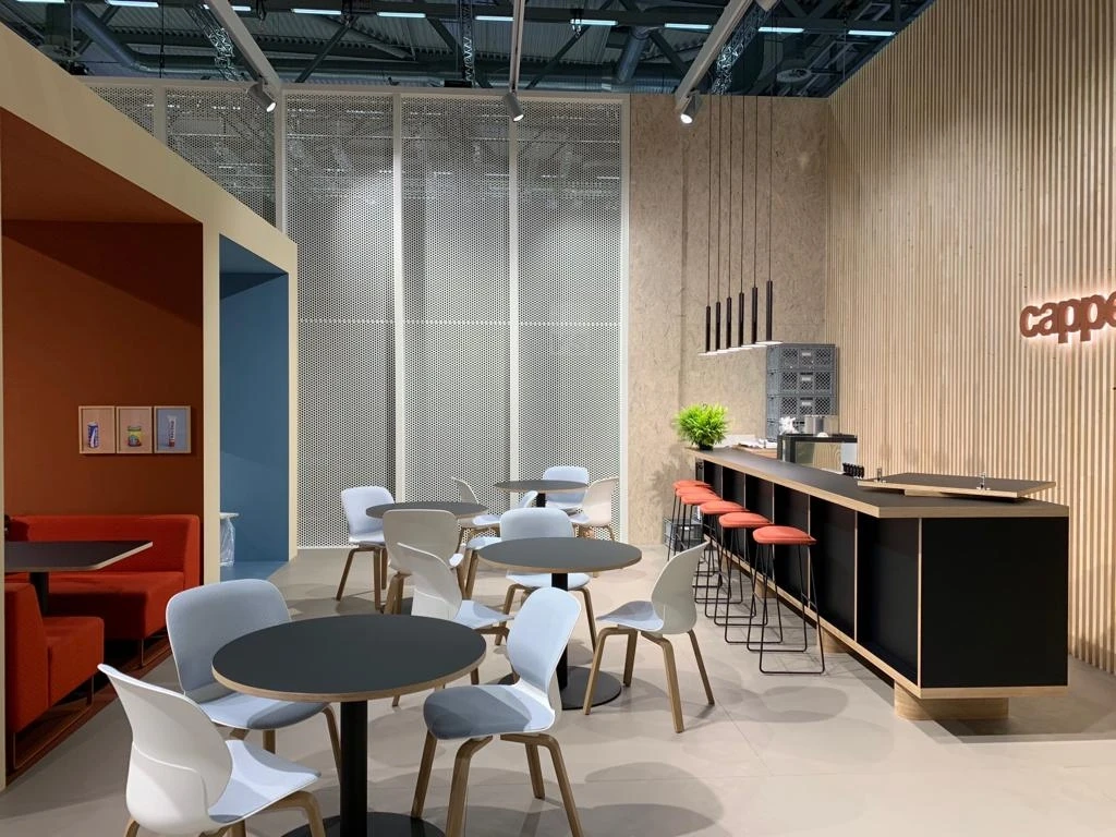 Coffee bar at the HAWORTH ORGATEC stand with black counter, coral bar stools, wooden slat wall with red cappellini neon and white café chairs.