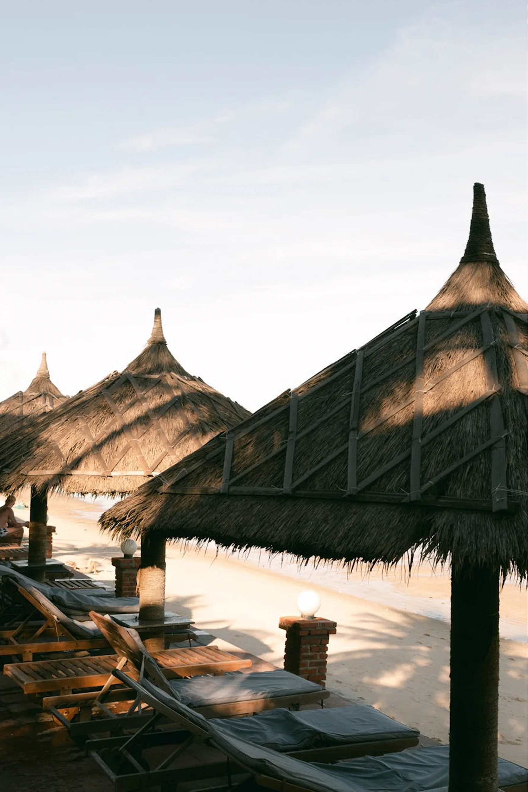 Thatched beachfront cabanas with lounge chairs beside the ocean.