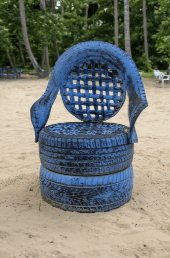 Blue chair made from repurposed car tires placed on a sandy beach with trees in the background.