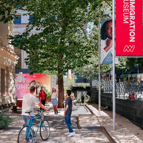 Människor promenerar och cyklar nära en trädbevuxen stig utanför Immigrationsmuseet, bredvid en röd och vit museibanner.