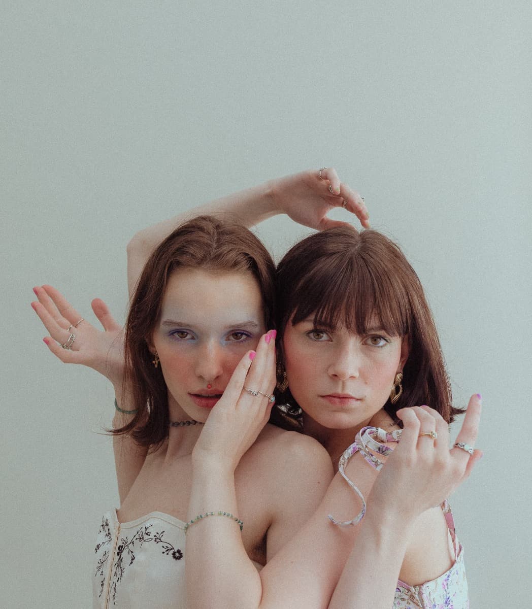 Models standing closely, one in a white corset and the other in a floral top, their fingers intertwined during a soft, ethereal photoshoot at Revelator Studio in Shreveport.