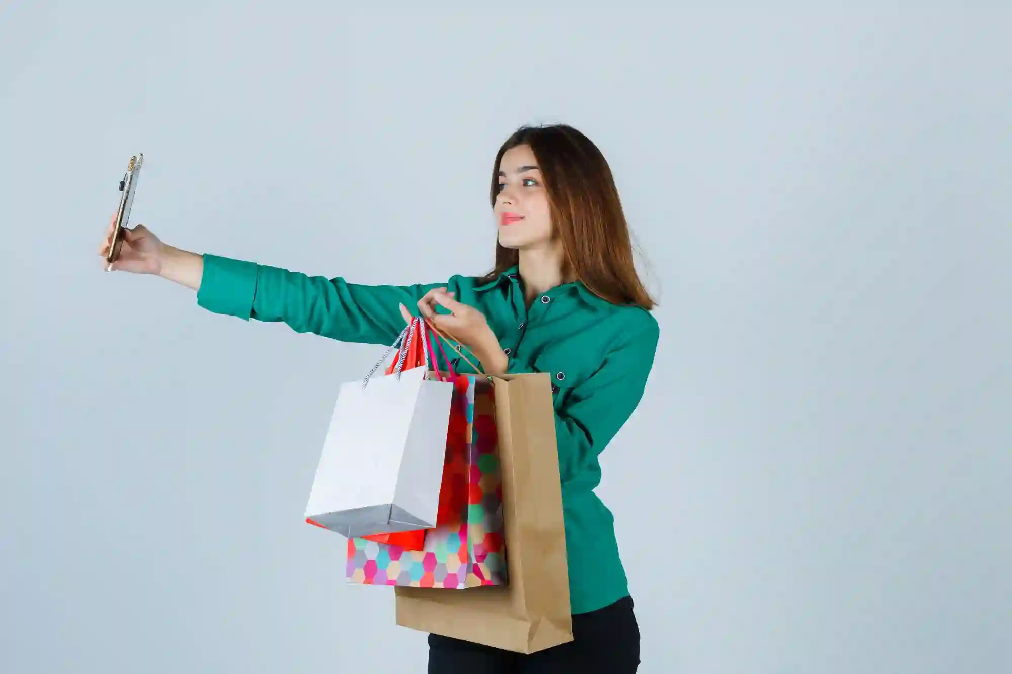 Young woman in a green shirt taking a selfie while holding several colorful shopping bags, celebrating a successful purchase.