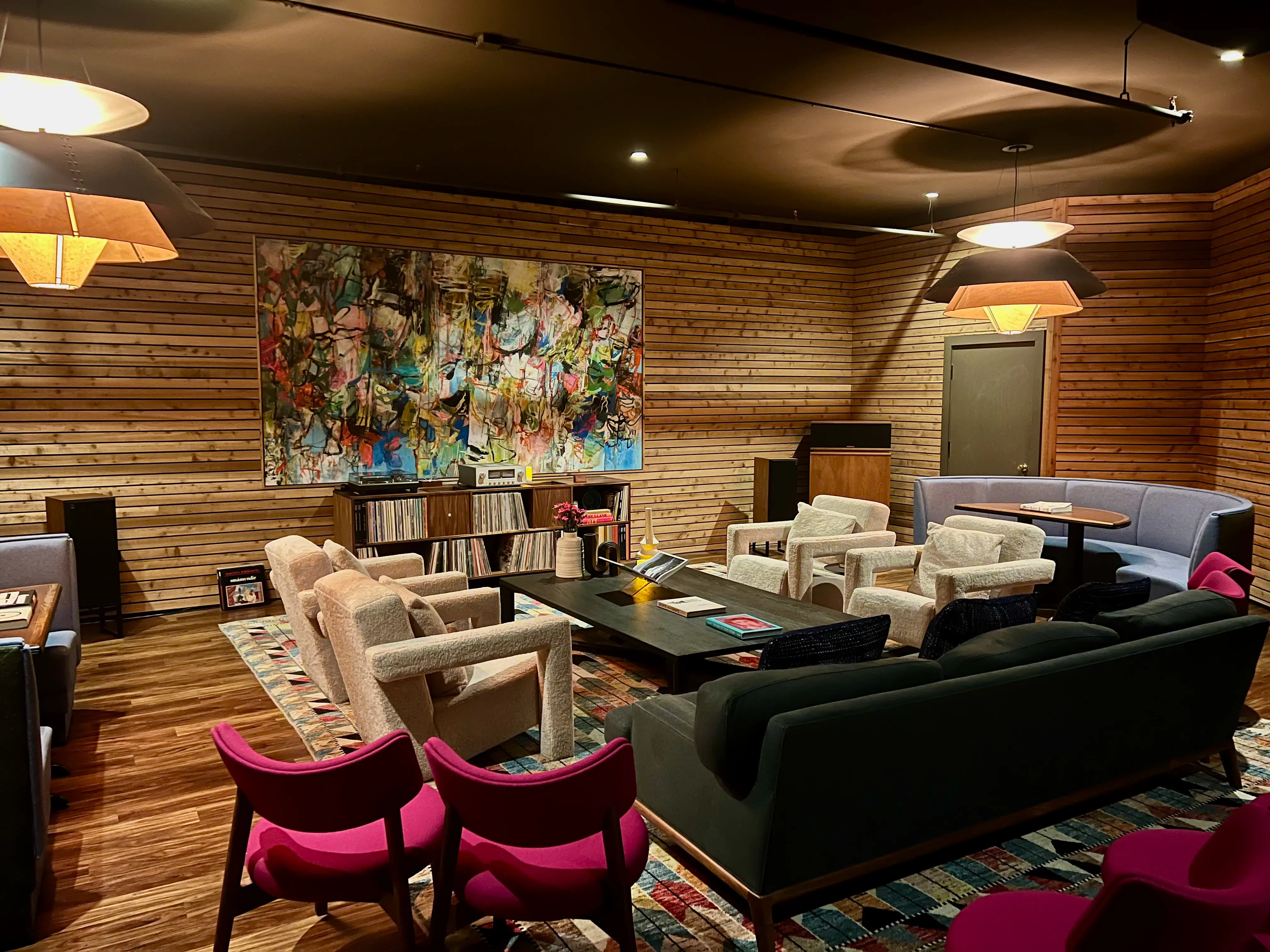 A cozy, modern lounge space at Shibuya HiFi in Seattle featuring wood‑slatted walls, comfortable seating arranged around low tables, vibrant chairs, and a large abstract artwork above a record collection, creating a stylish setting for the HiFi Room Experience.