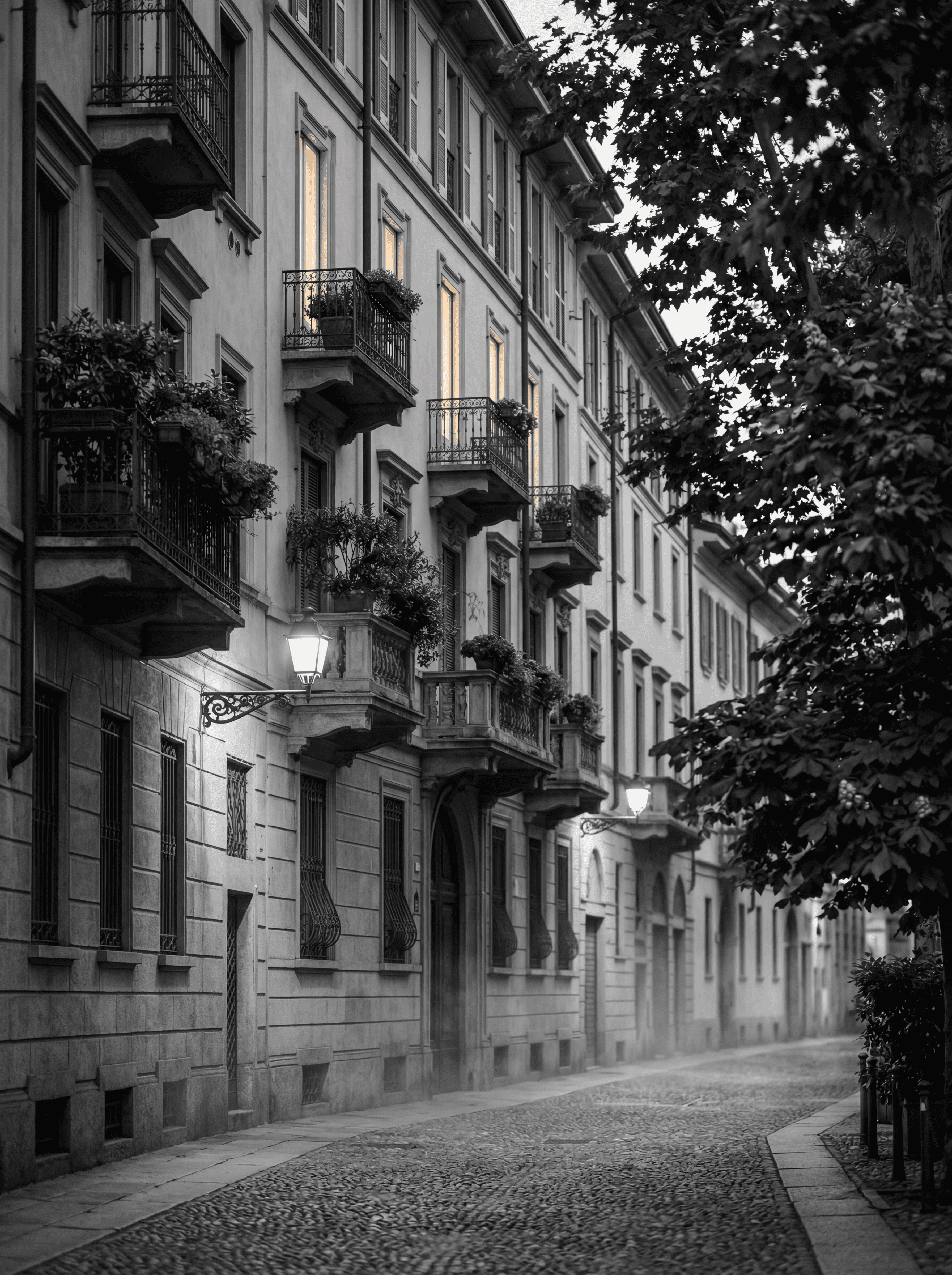Brera district in central Milan at dusk with classic Italian palazzi and illuminated upper-floor windows, illustrating the London-to-Milan wealth migration corridor, monochrome editorial photograph