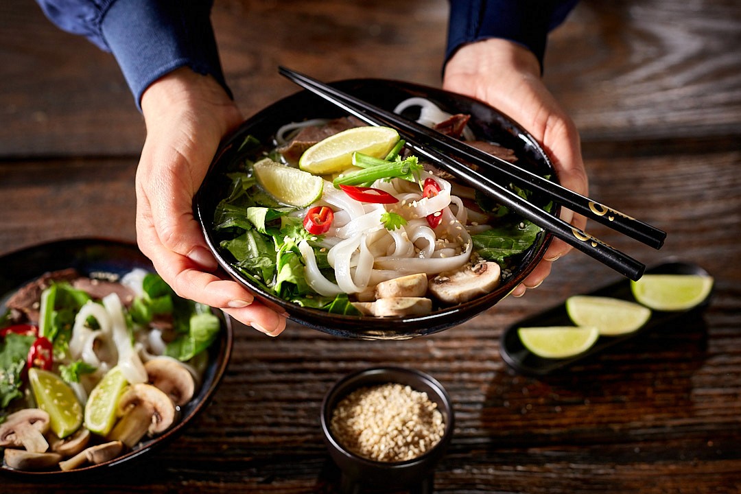 Two hands hold a bowl of fresh pho, garnished with lime wedges, red chili slices, cilantro, and mushrooms, with a pair of chopsticks resting on the rim, alongside a small bowl of sesame seeds on a wooden table.
