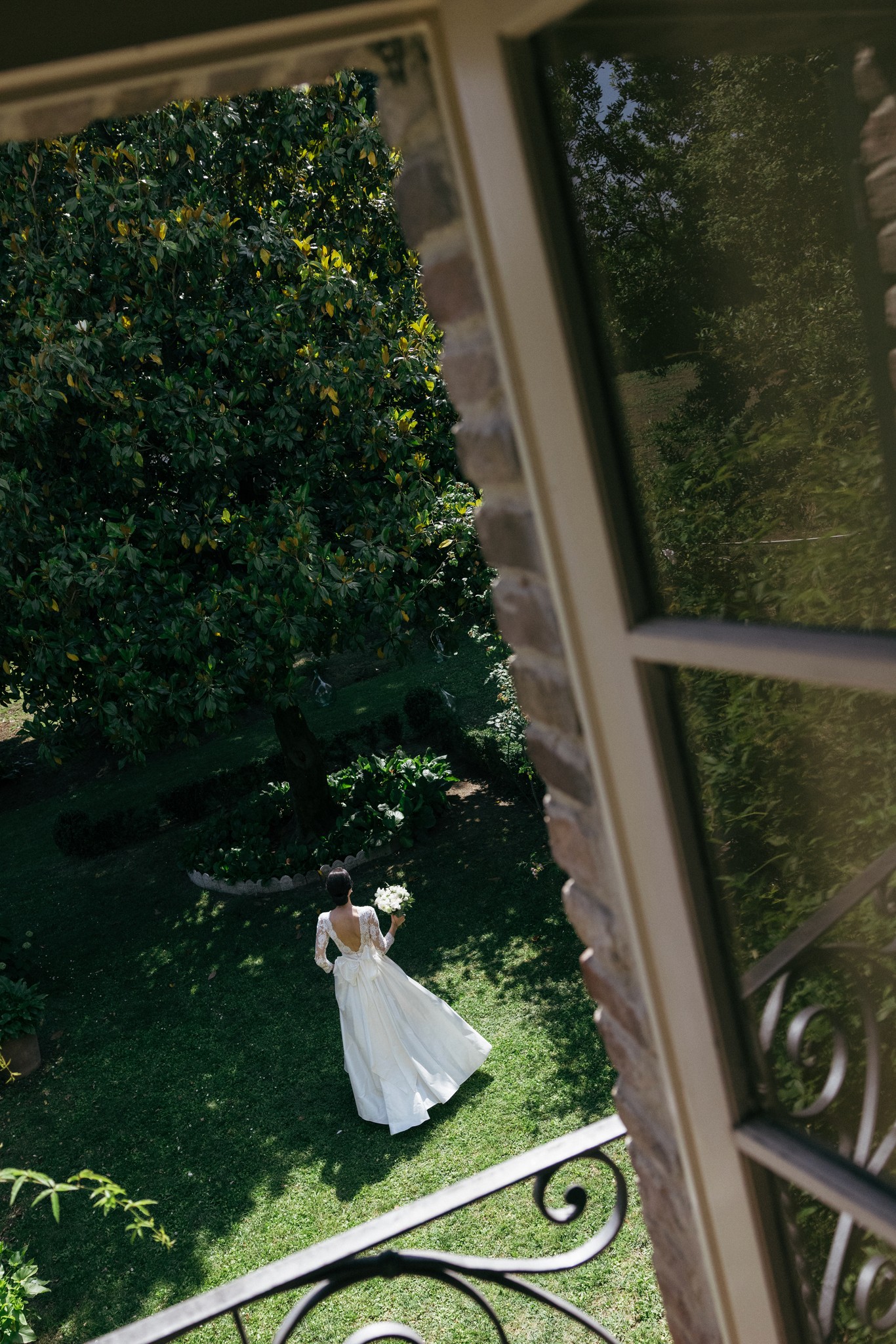 Sposa in abito bianco nel giardino della villa, vista da finestra con persiane verdi, luce naturale tra la vegetazione
