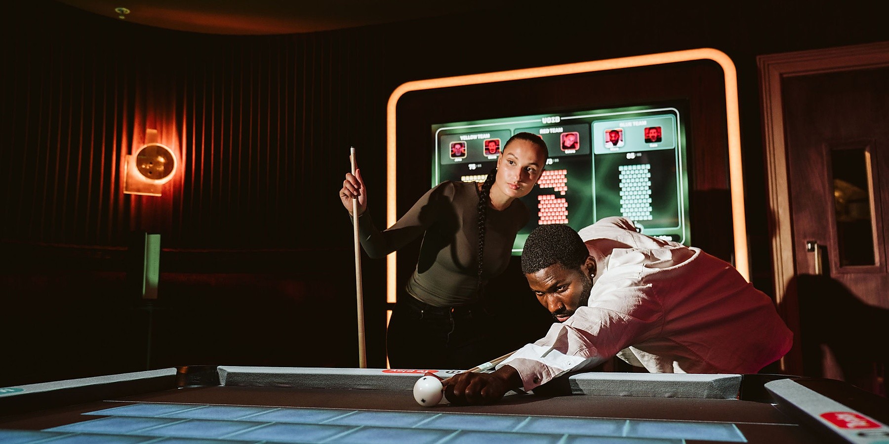 wo guests playing on a digitally enhanced pool table inside a warmly lit social entertainment venue.