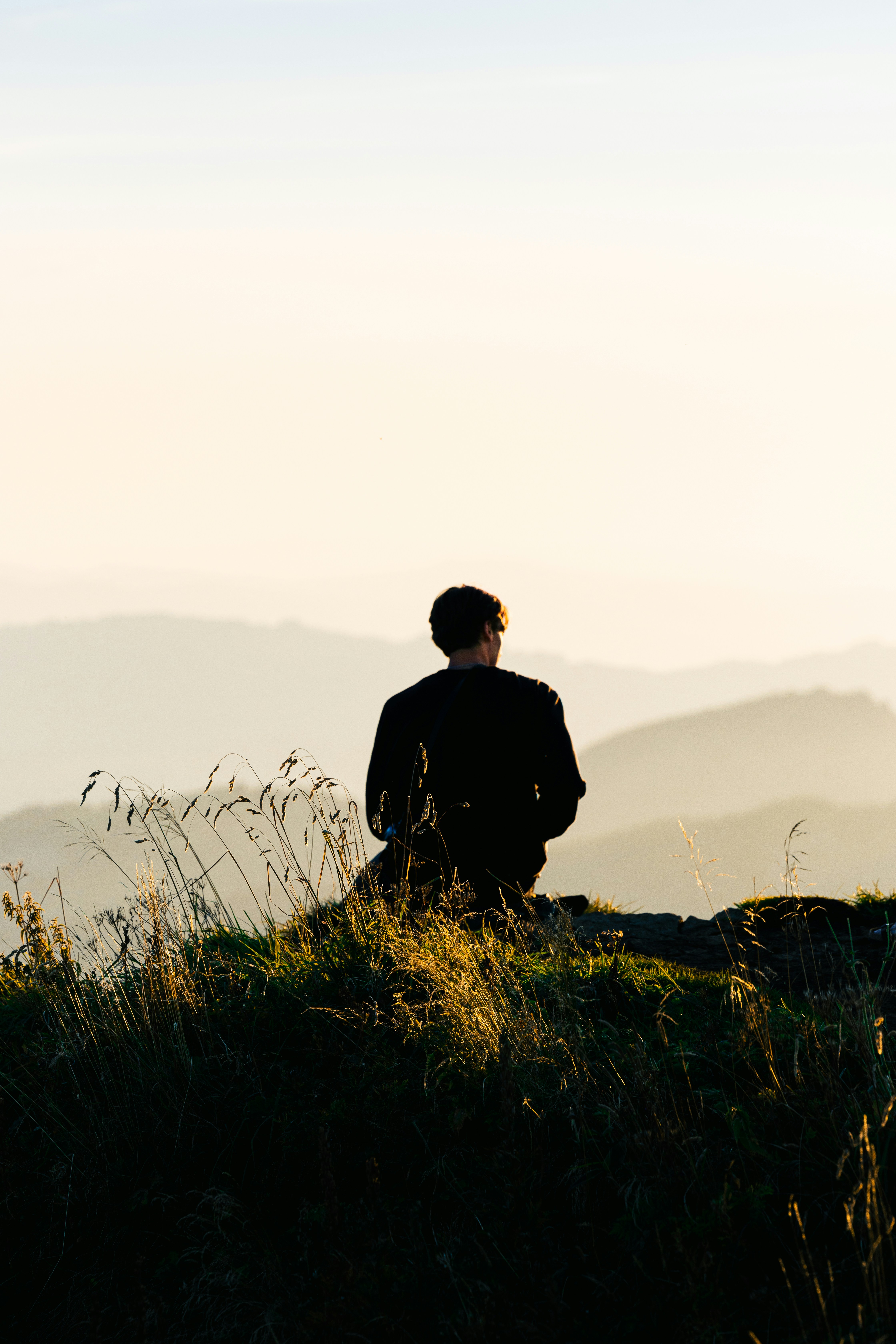 Person sitting on grassy hilltop overlooking mountains at sunset.