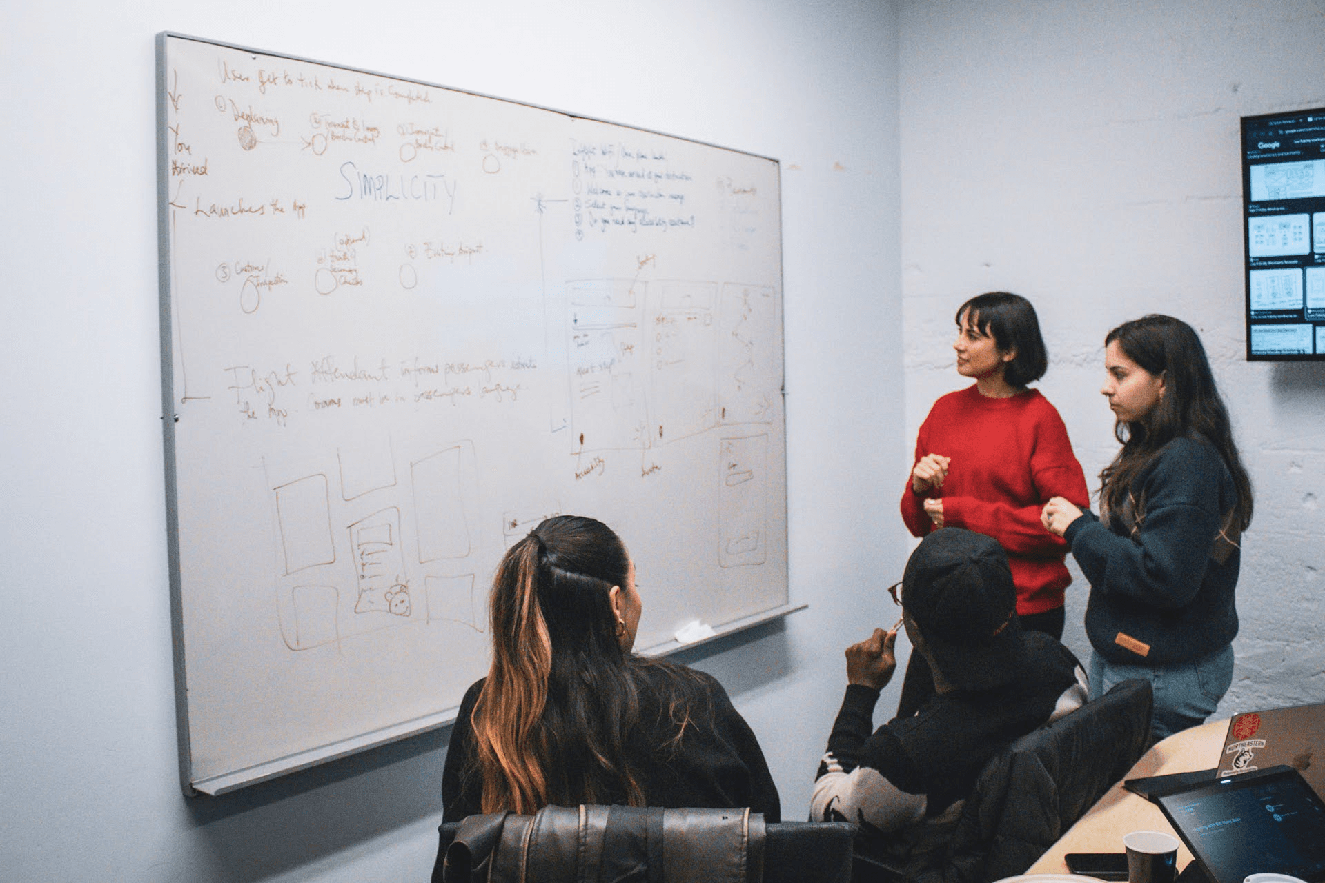 Close-up of two professional women in conversation in a contemporary office space. The woman facing the camera has long wavy brown hair, wears a burgundy knit top and nose ring, and displays an engaged, friendly expression. Another woman with short dark hair and pearl earrings is shown from behind. The bright, modern workspace features other colleagues working at desks in the background.