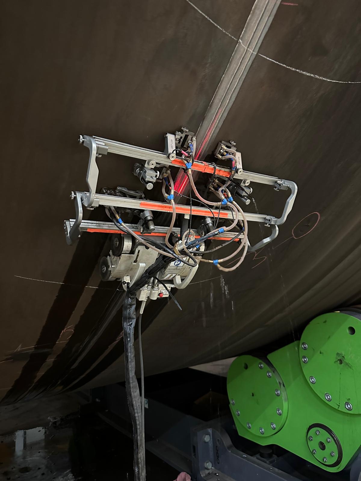 A device is mounted on a large, dark metal surface, with cables and hoses visible. Nearby, a green circular component is partially visible. The scene conveys industrial precision.