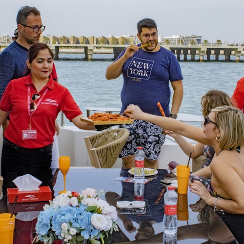一班人在船上用餐，兩位穿紅色衫的侍應，一位穿藍色T恤的男士，以及三位坐在那裡的人。