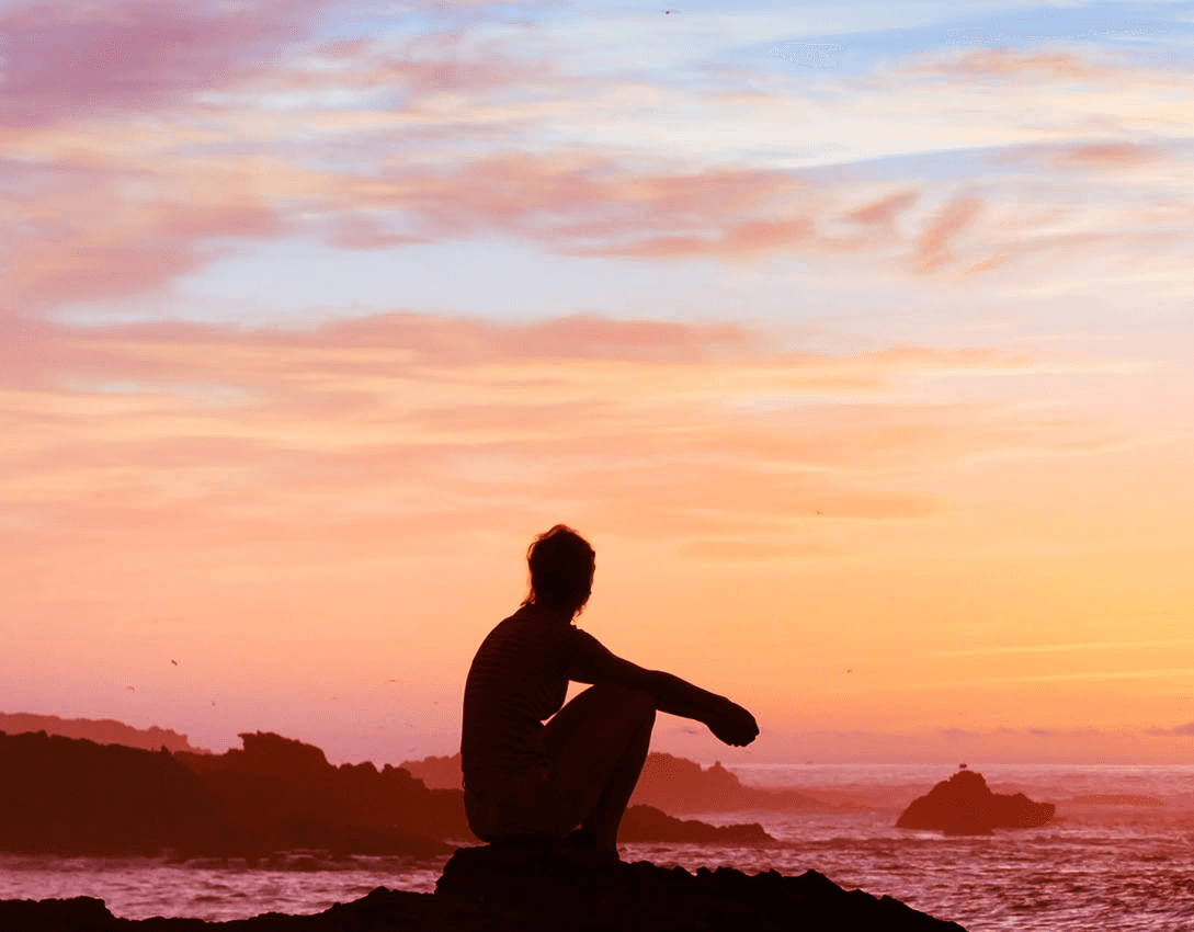 A lady perched on a rock by the sea, gazing at the setting sun