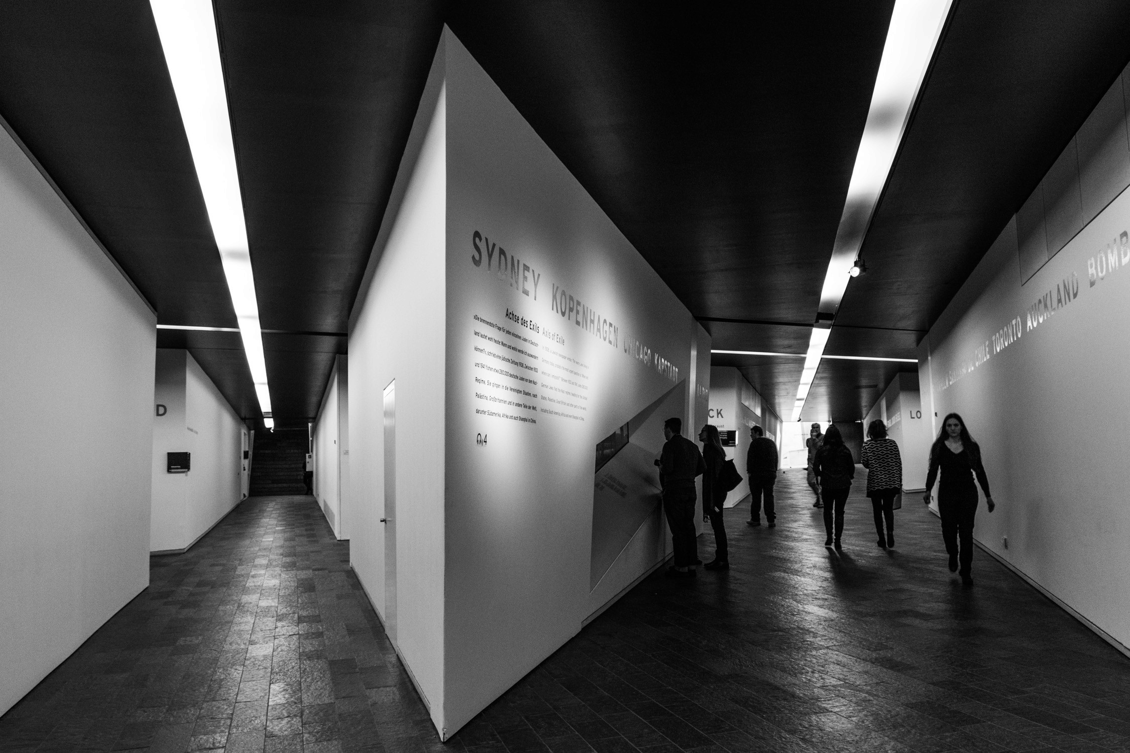 People walk through a modern underground corridor with white walls, dark ceilings, and long linear light fixtures.