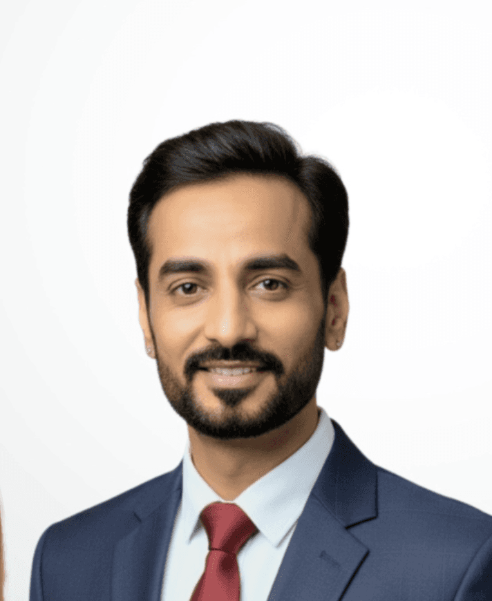 A smiling man with curly hair is sitting at a desk, looking at the camera, with a laptop and a lamp in the background.