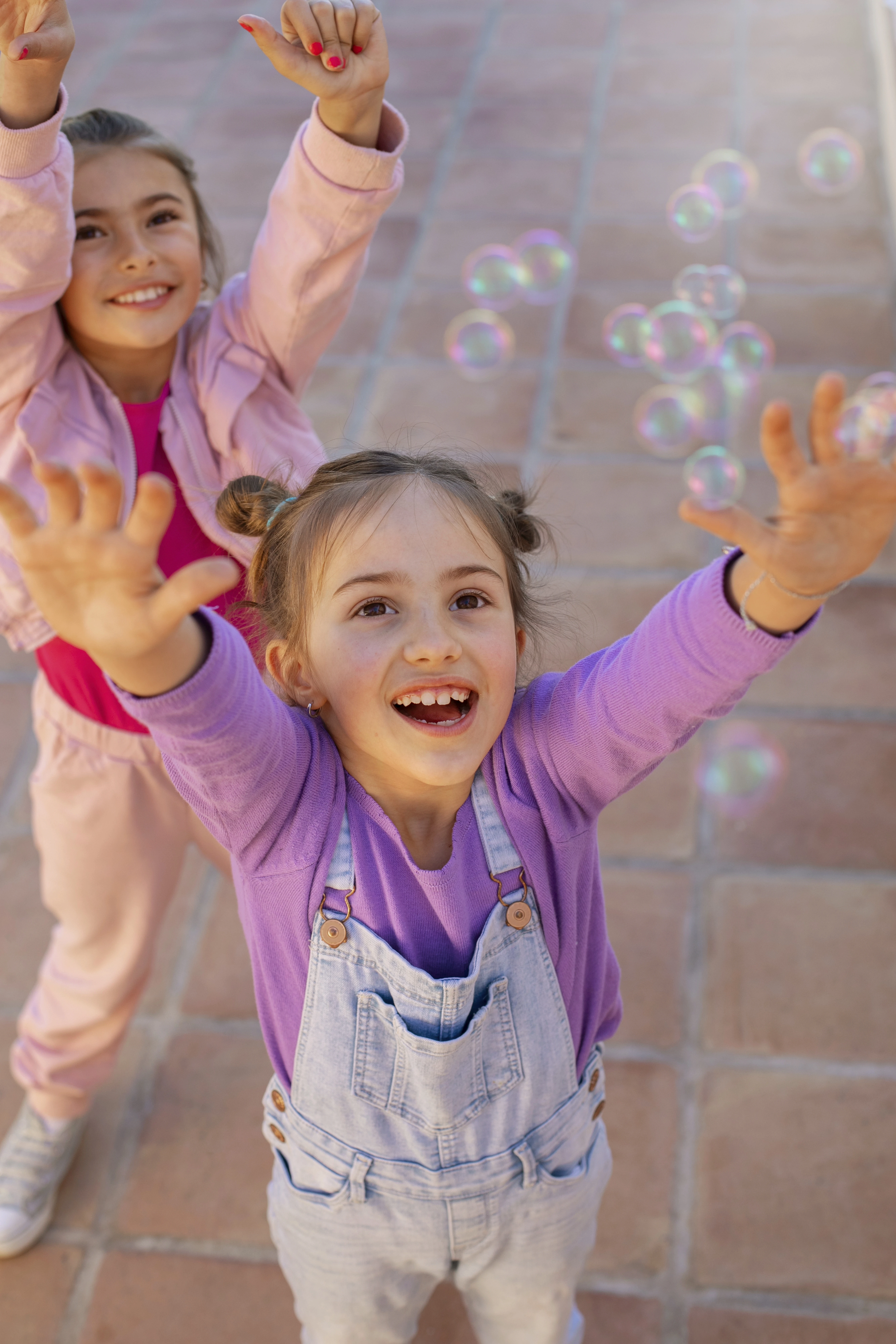 Duas meninas sorridentes com os braços levantados tentando alcançar bolhas de sabão que flutuam no ar em um ambiente externo ensolarado.