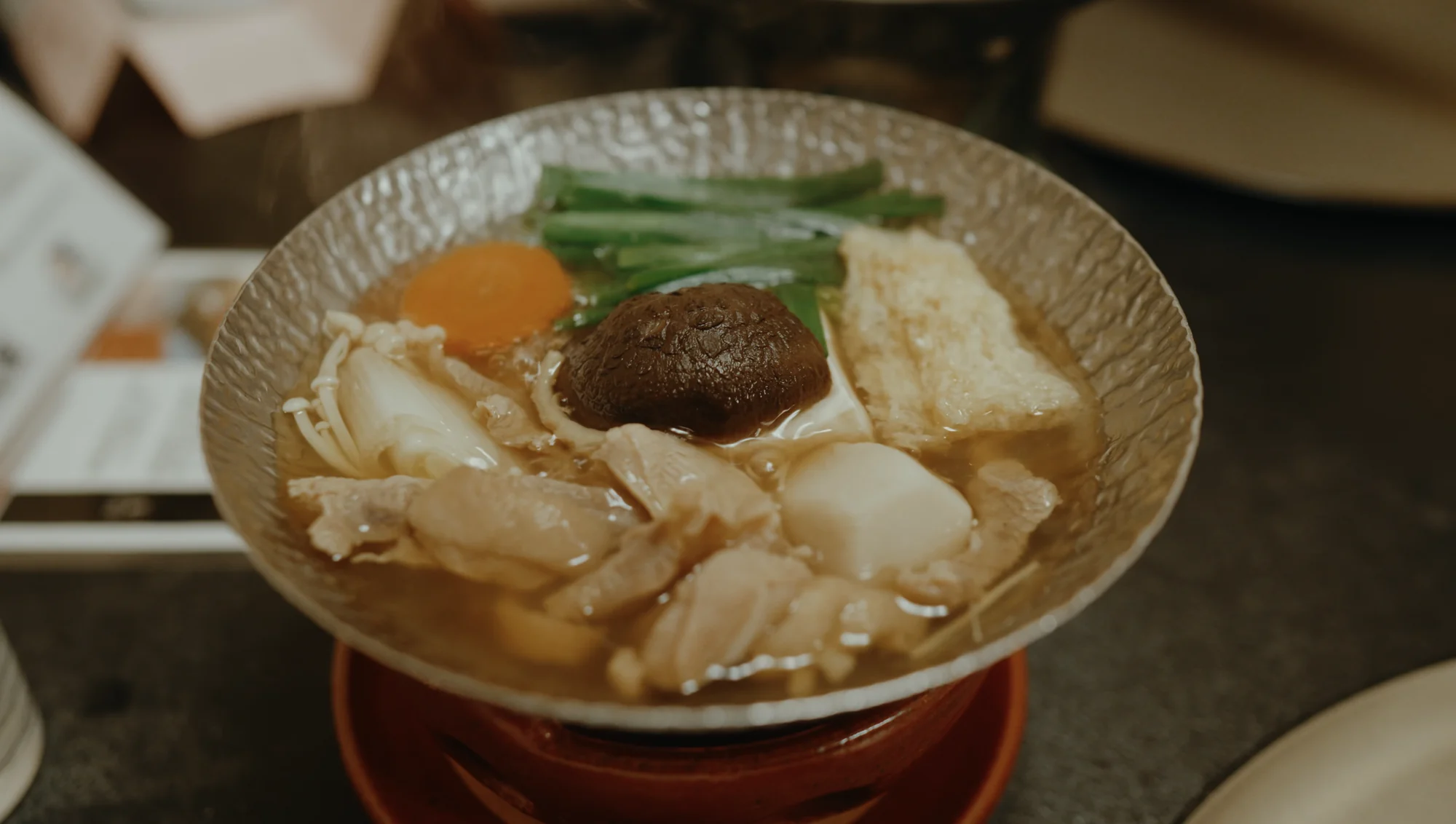 A clear hot pot of chicken, tofu, shiitake and greens on a red lacquer stand