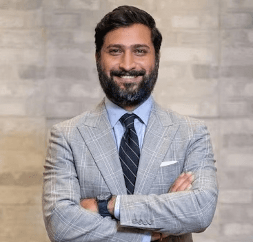 Portrait of Neeraj, wearing a dark-colored kurta with arms crossed, looking confident against a light purple gradient background.
