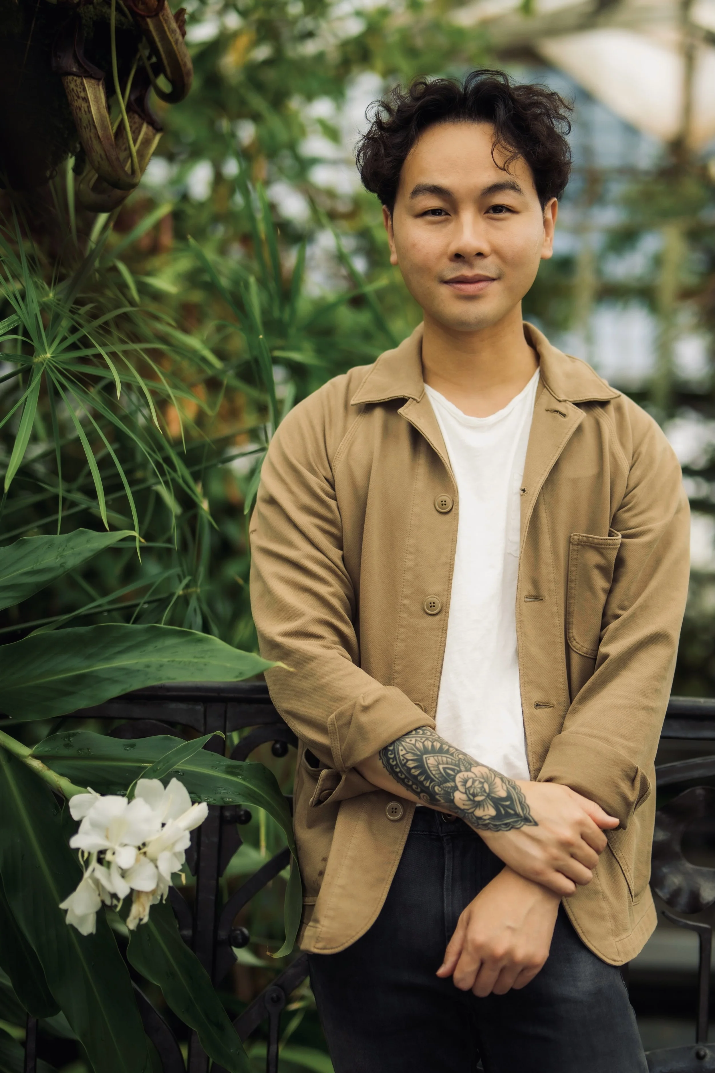 Uma pessoa com cabelo cacheado e uma tatuagem em um dos braços está em um exuberante jardim verde, usando uma jaqueta bege sobre uma camisa branca, com flores brancas desabrochando por perto, transmitindo uma atmosfera tranquila e natural.