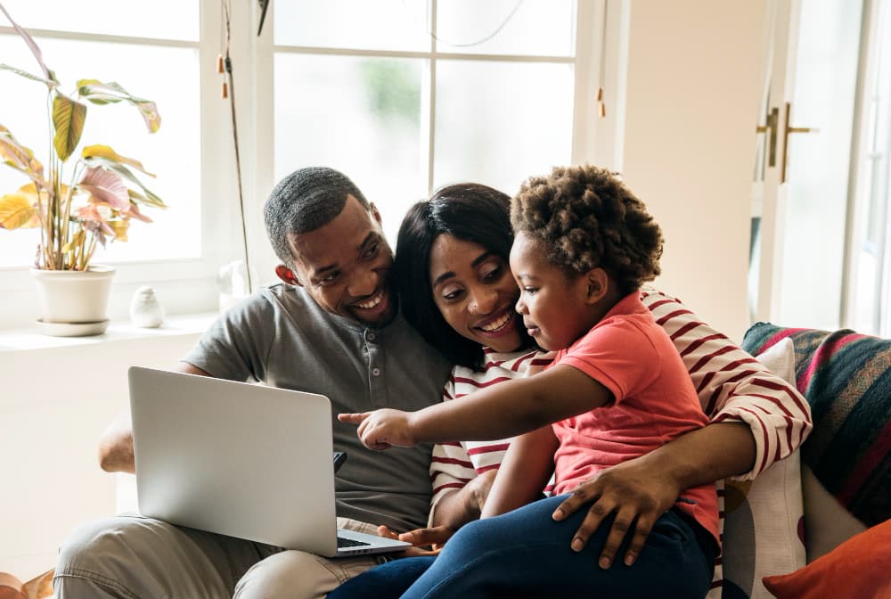 A family sits together on a couch at home, smiling as they look at a laptop with their young child. The image represents learning about home loan options and reflects the supportive, family-focused approach of Chris Lewis Home Loans.
