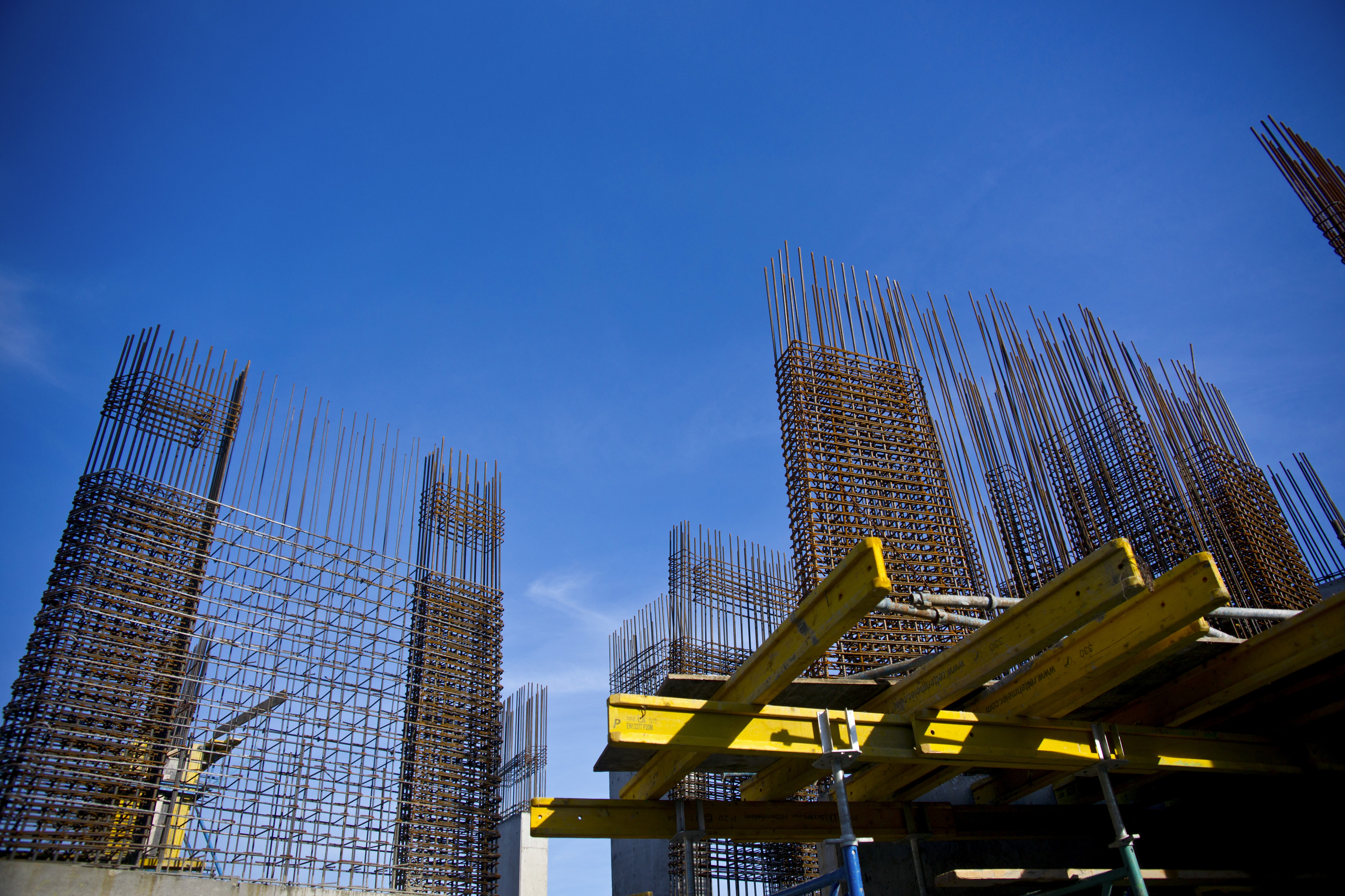 Steel rebar structures rise against a vivid blue sky, part of a construction site. Yellow scaffolding adds contrast, conveying growth and progress.
