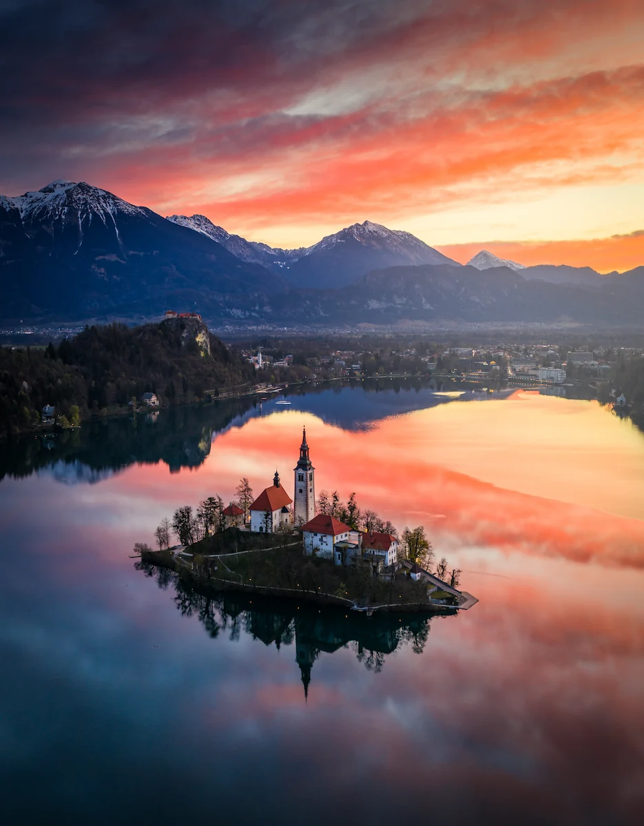 Lake Bled island church at sunrise, surrounded by still water reflecting a bright orange and pink sky, set against the Julian Alps in Slovenia.