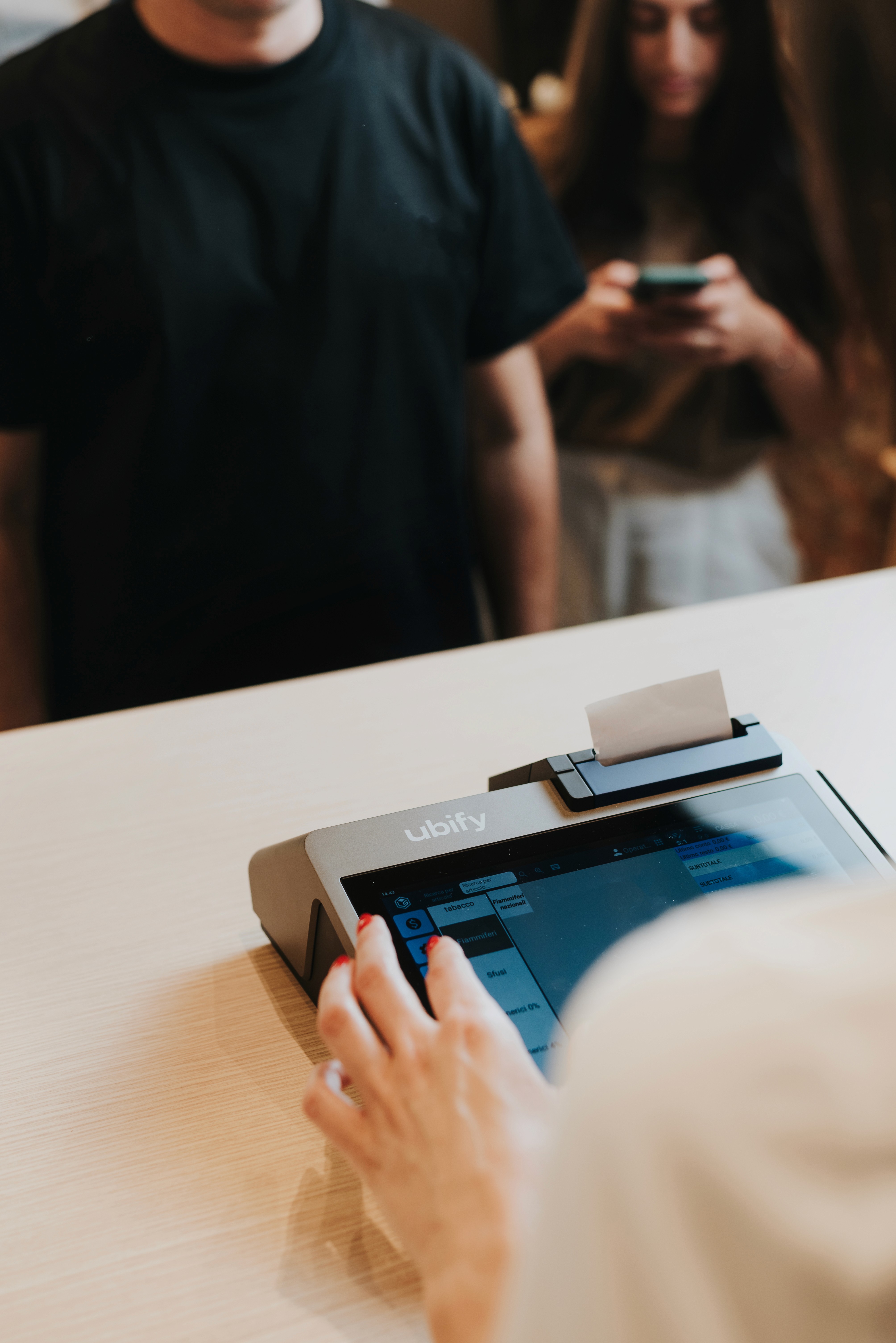 Commerciante utilizza il touchscreen della Cassa Ubify mentre due clienti attendono al bancone.