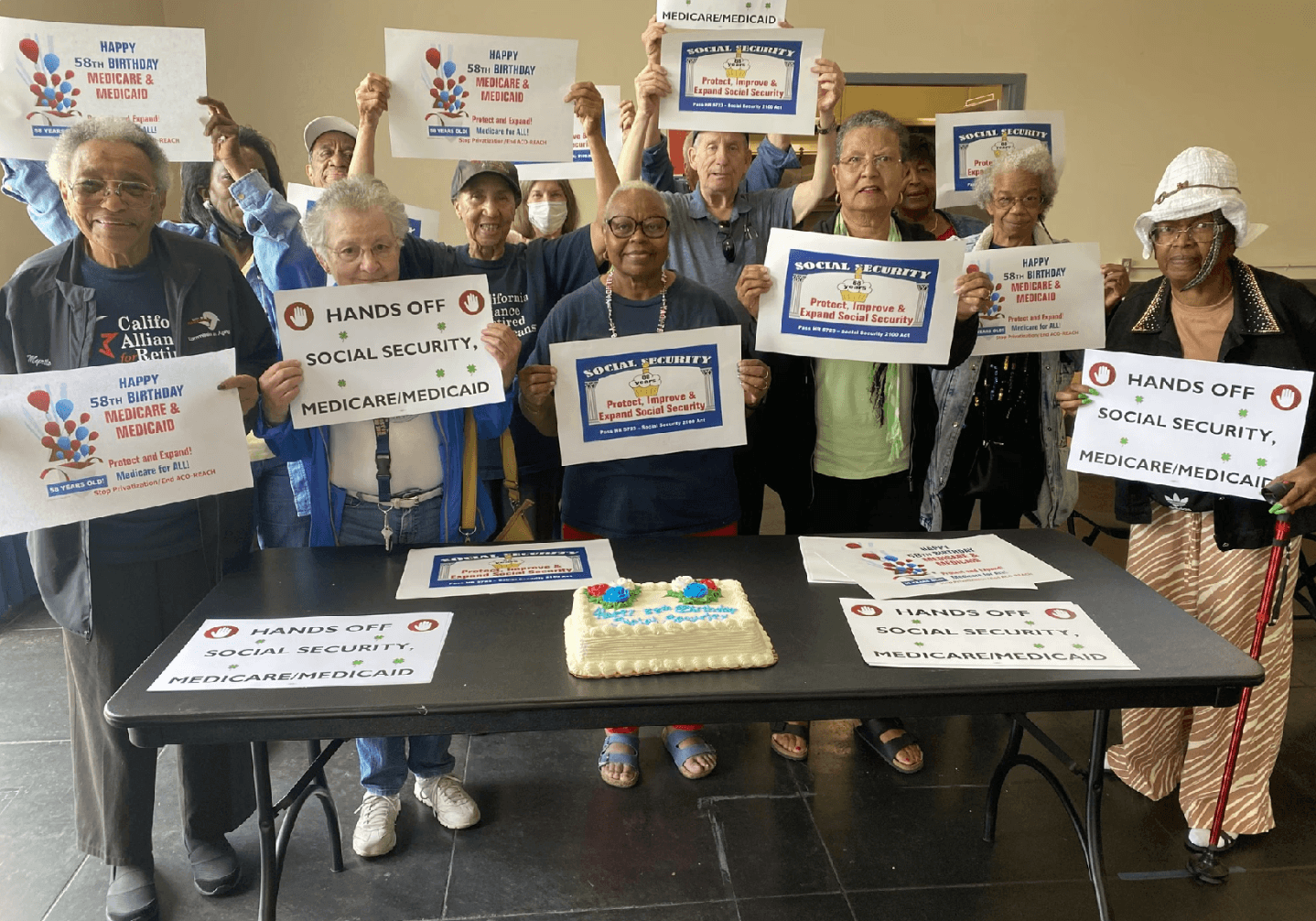 A group of older adults stands behind a table with a decorated cake, holding signs that advocate for protecting Social Security, Medicare, and Medicaid. The mood appears celebratory and united, with messages like “HANDS OFF Social Security, Medicare/Medicaid” and “Happy 58th Birthday Medicare & Medicaid.”