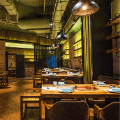 Una vista interior de un restaurante con mesas y sillas de madera, con una iluminación suave. El ambiente se ve acogedor y elegante, con una decoración mínima y una atmósfera cálida.