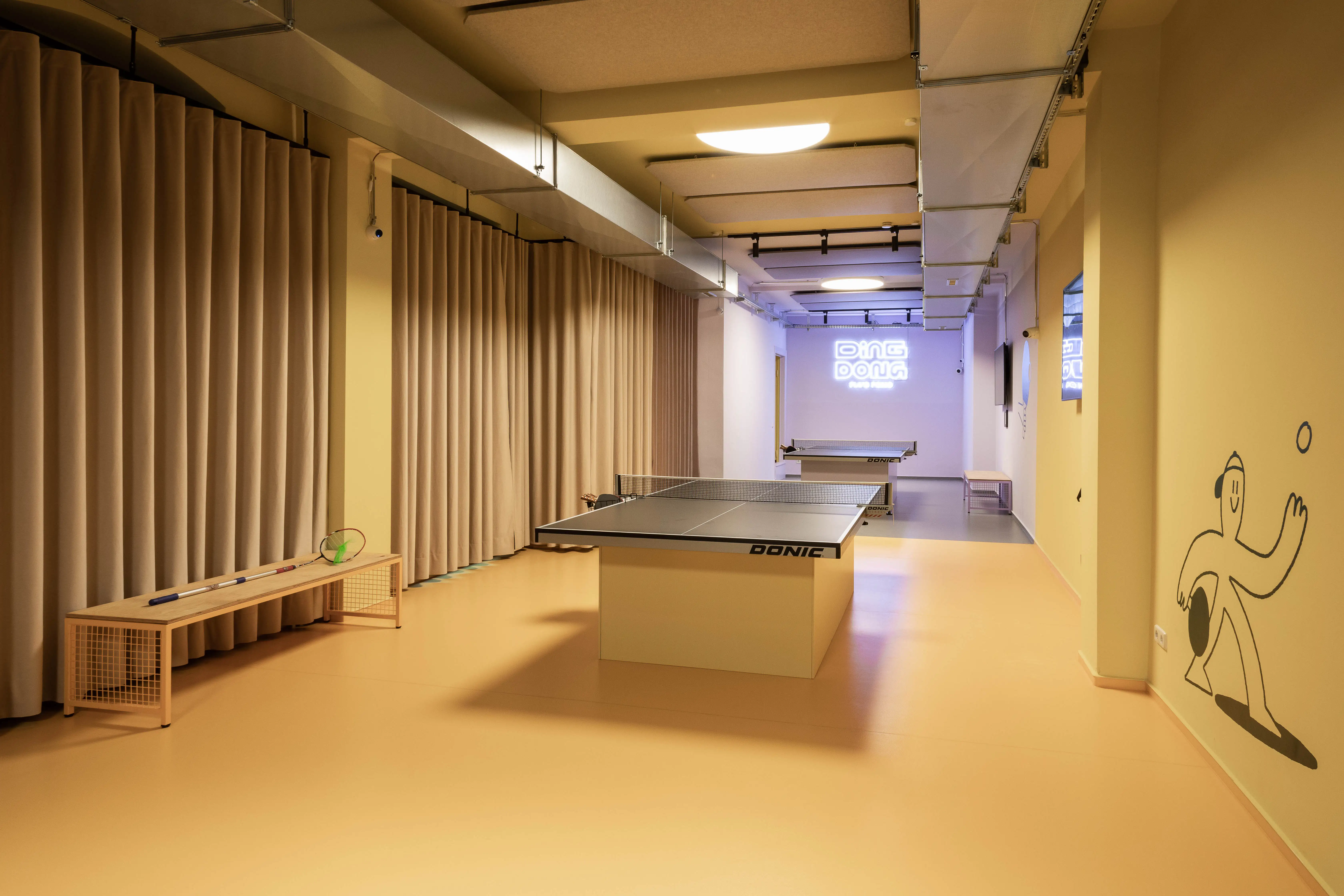 Play hall at DING DONG PING PONG with table tennis tables on a peach-colored floor, beige curtains, a player mural and a glowing logo sign at the far end.