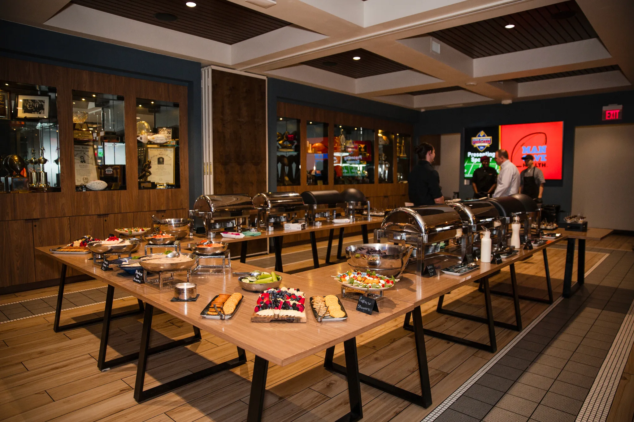 A modern restaurant banquet room features wooden tables laden with chafing dishes, appetizers, and desserts, while staff members attend to guests near a red digital display and wood-paneled walls with trophy cases.
