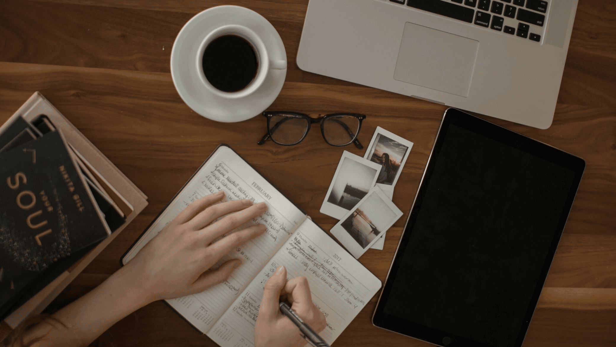 A person sits at a wooden desk writing in a notebook. There is a laptop, coffee cup, glasses, a stack of books, and a tablet on the desk.