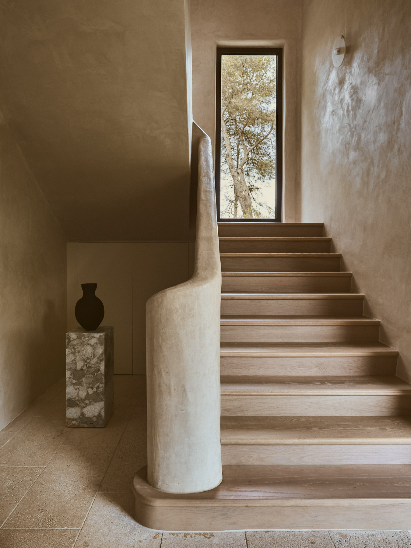 Sculptural staircase illuminated by a vertical window in a contemporary Mediterranean home.