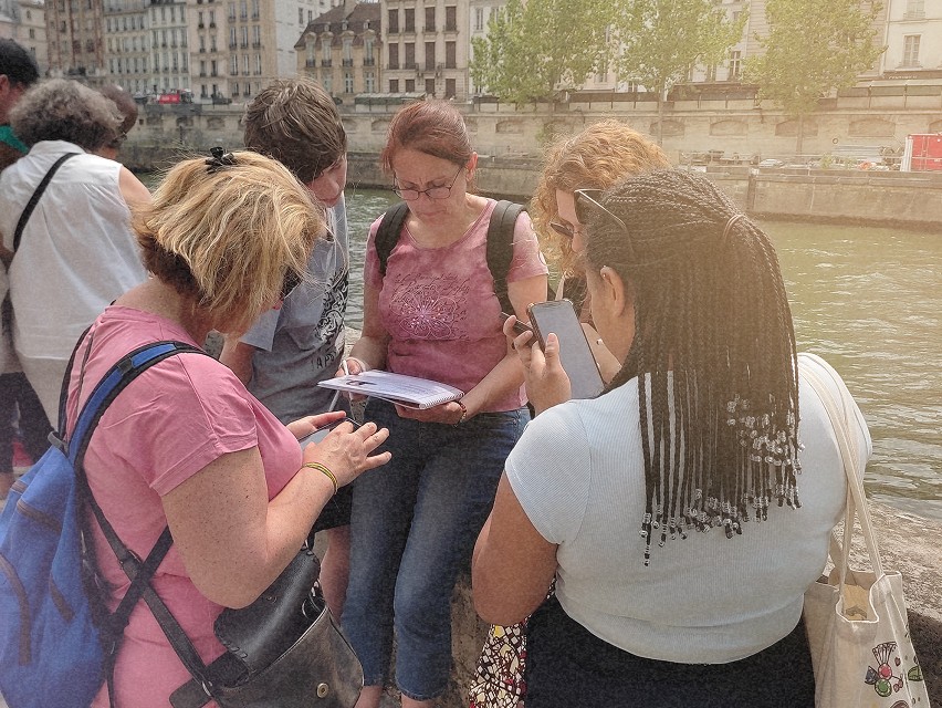 Un groupe de personnes, debout près de l’eau, lisent et consultent leurs téléphones, avec un bâtiment en arrière-plan.