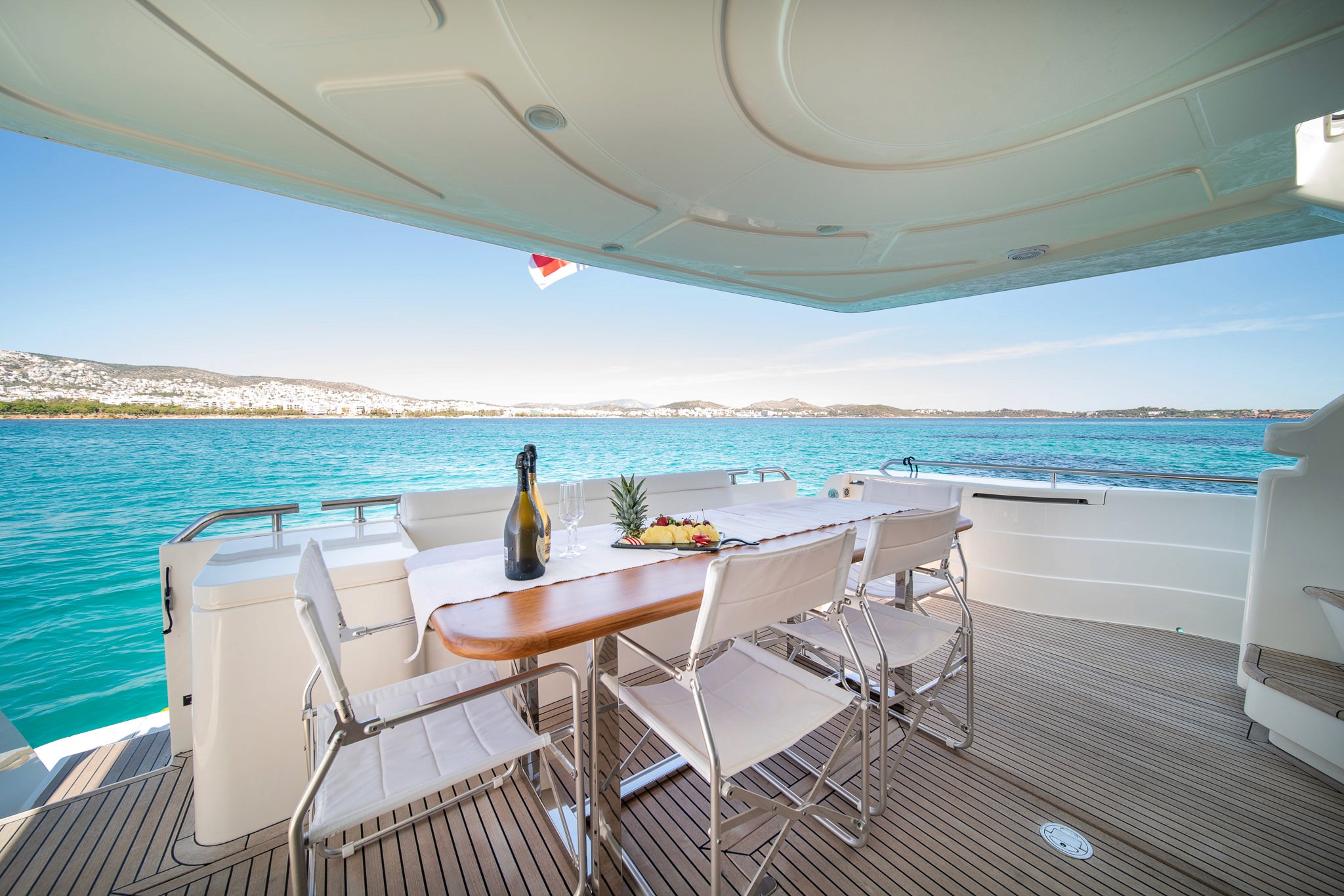 Elegant dining setup on the aft deck of Ferretti 68 yacht with white chairs around wooden table, overlooking turquoise Aegean waters and Greek coastline.