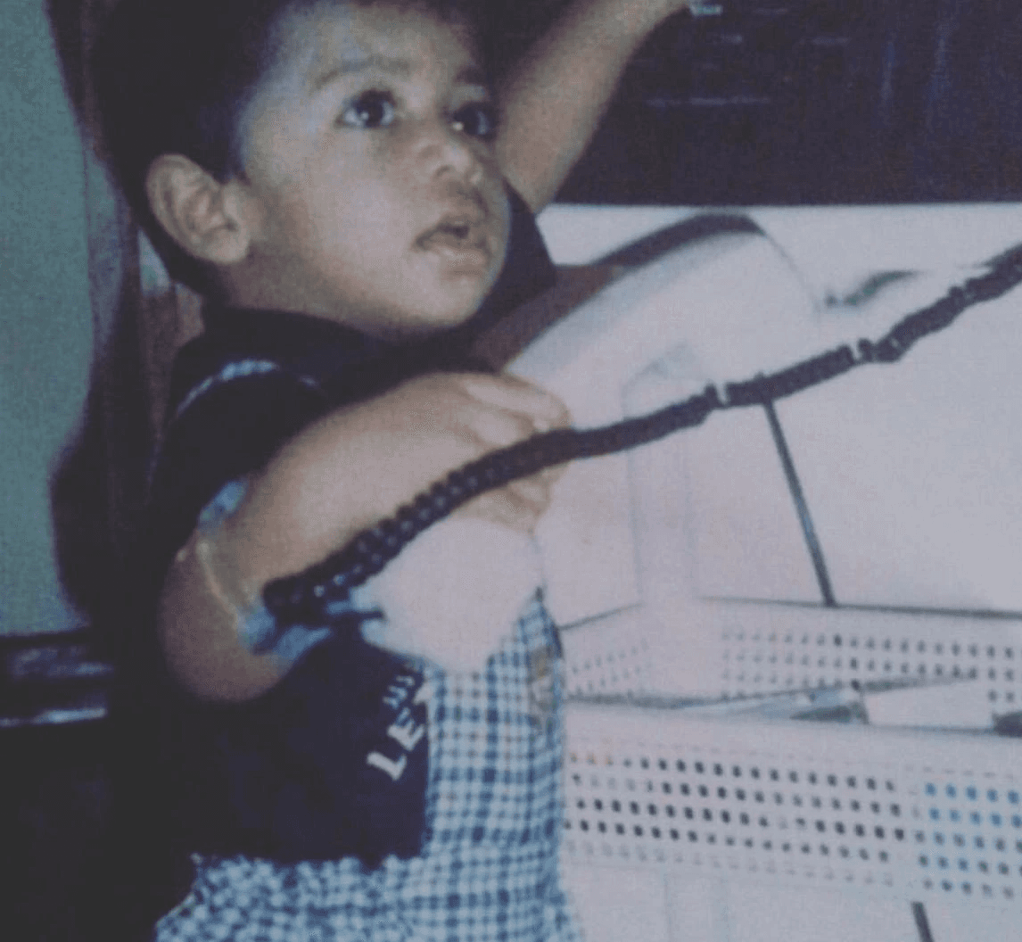 Portrait of a smiling woman with natural curly hair wearing a pale yellow T-shirt and gold hoop earrings against a gray background.