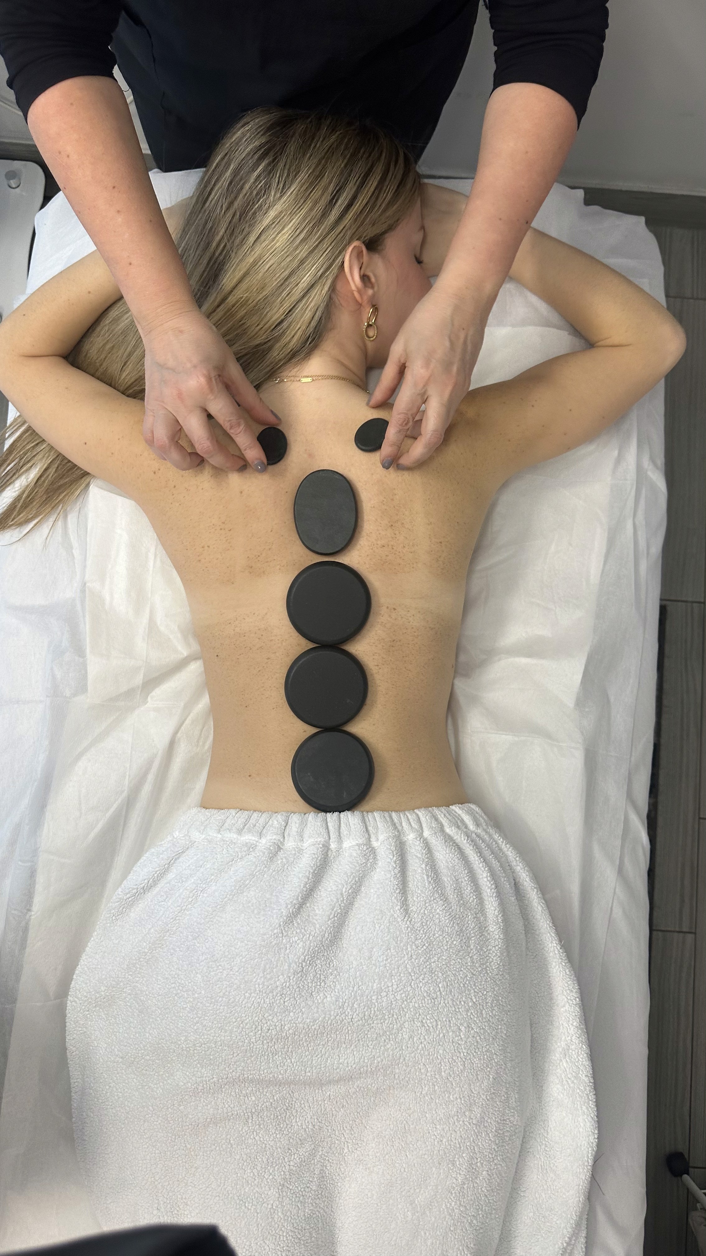 A cozy and inviting spa room with a neatly made bed, soft lighting, and warm wooden tones. The scene is partially framed by green plants, adding a natural and soothing touch.