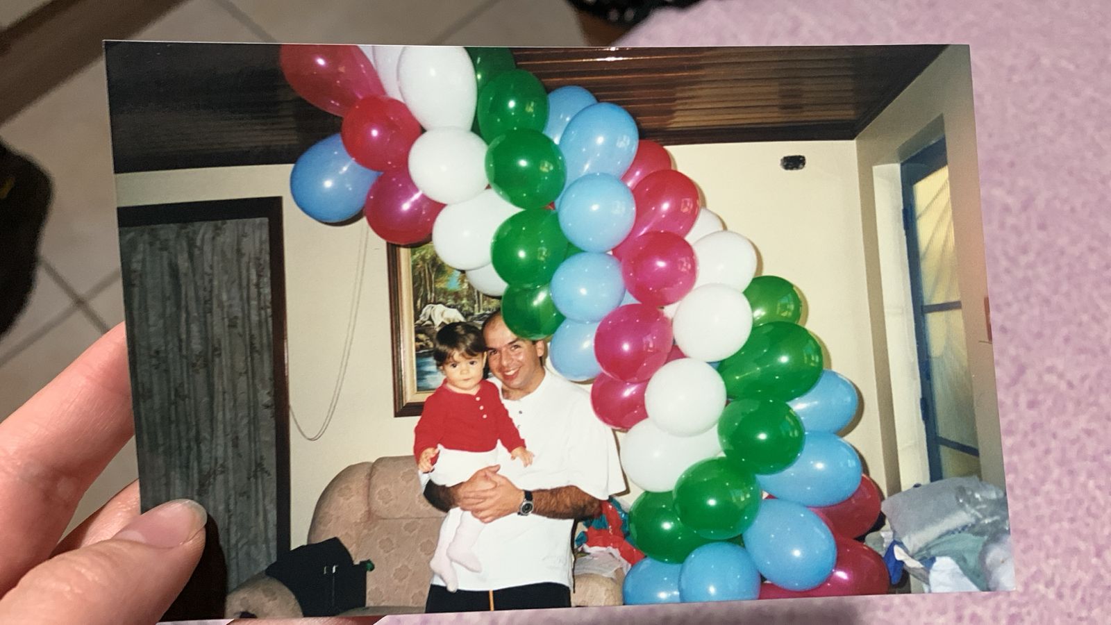 Pai segurando filha no colo com arco de balões no fundo