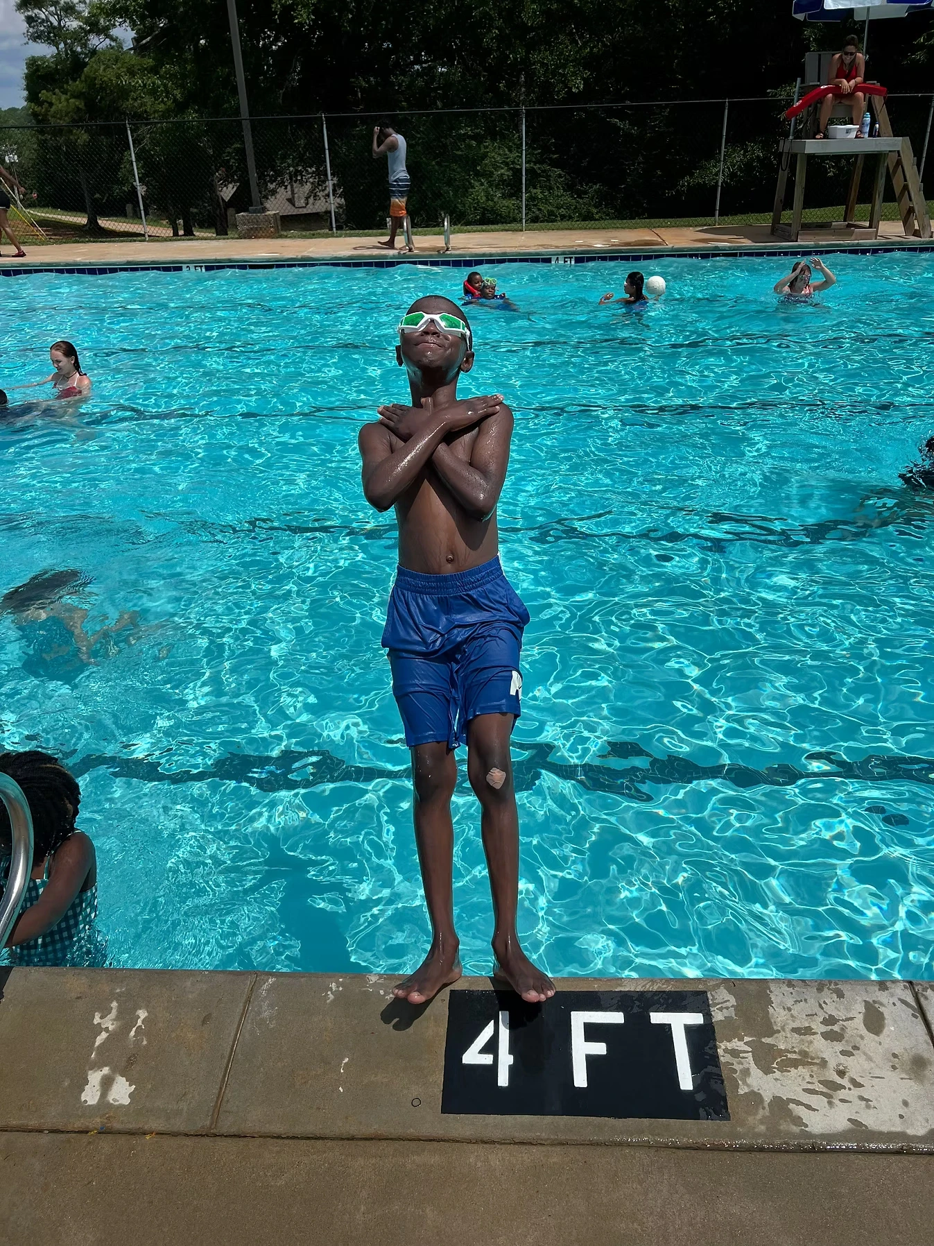 A young boy wearing swim goggles and blue swim trunks stands at the edge of a swimming pool with his arms crossed, about to jump in.