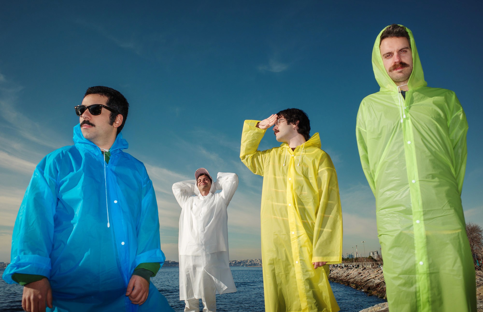 Four individuals stand near a rocky shoreline, wearing brightly colored raincoats in blue, white, yellow, and lime green, under a clear blue sky.