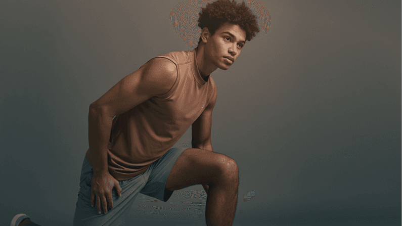 A young man in athletic wear kneels on one knee, poised and focused, against a neutral backdrop.