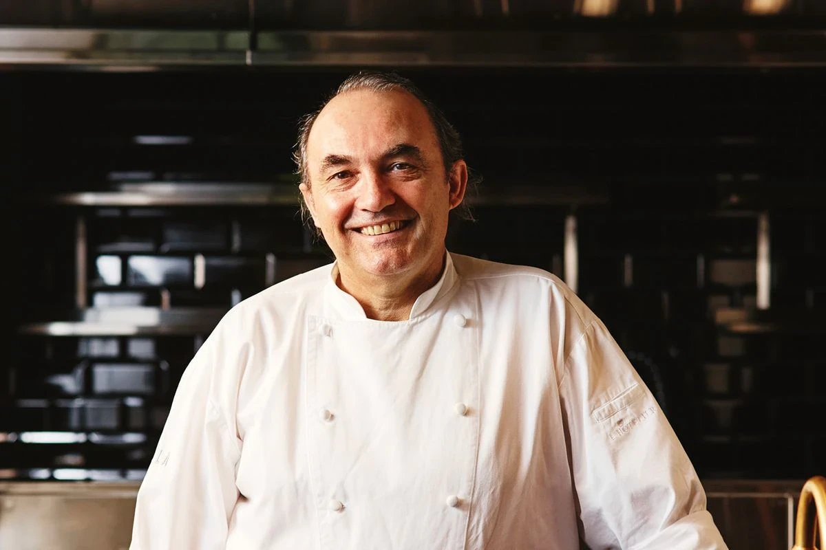 Smiling chef wearing a white coat stands in a kitchen.