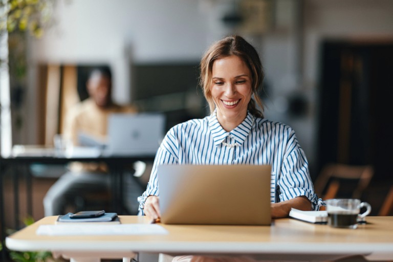 A woman in a striped blouse smiling while working on a laptop in a café or modern workspace with a coffee cup nearby