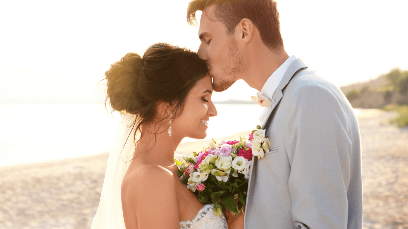 A couple in wedding attire embraces on a beach, sharing a tender moment under the sunlight.
