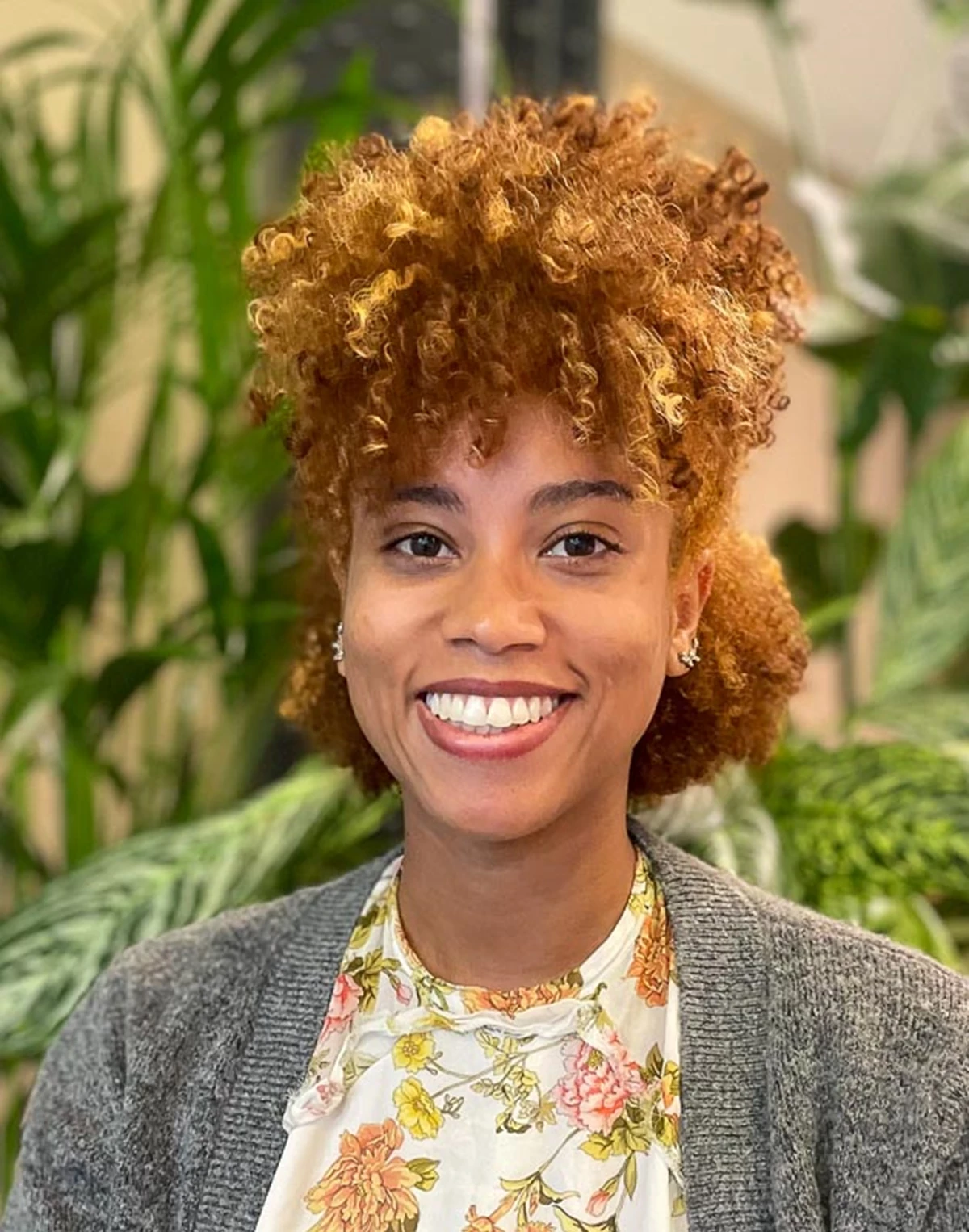 A woman with auburn natural curly hair is smiling at the camera wearing a floral shirt and a grey cardigan, in the background are lots of green plants